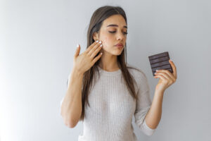woman holding a chocolate bar while touching her face and wondering "does sugar cause acne?"