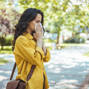 woman blowing her nose or sneezing into a tissue in the park while experiencing tree pollen allergies