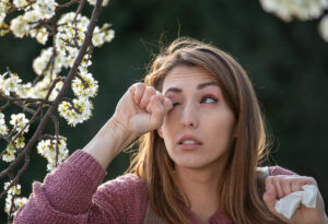 Young pretty girl rubbing eyes beside blooming tree in spring time. Itchy eyes from allergy