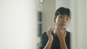 Asian teenage boy washing face skin by foam cleanser in front of bathroom mirror.