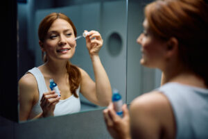 Smiling woman using face serum while taking care of her skin in front of a mirror.