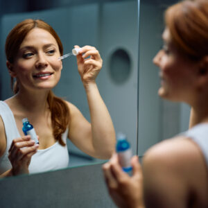 Smiling woman using face serum while taking care of her skin in front of a mirror.