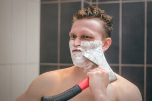 Frown man with brutal face is shaving with axe shaver in the bathroom.