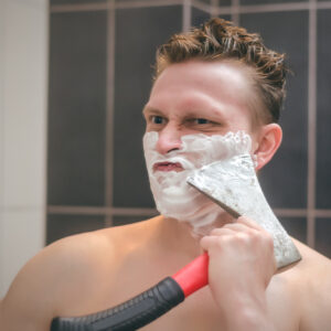 Frown man with brutal face is shaving with axe shaver in the bathroom.