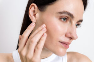 A young woman applies moisturizing face cream on skin, using skincare products, highlighting her clear and smooth complexion. She focuses on beauty treatments that enhance natural health